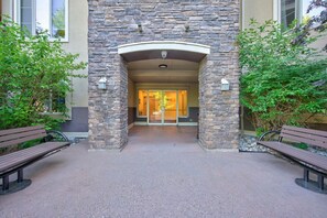 A stone entrance with benches, a glass door, and a building with a beige facade. - Mountain View 3 Bd | Pool, Hot Tub & Golf 2208 (Kelowna)