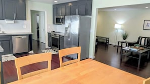A modern kitchen with stainless steel appliances, a dining area with a wooden table and chairs, and a living room with a sofa and floor lamp.