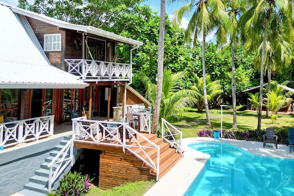 Une maison en bois à deux étages, dotée d’un balcon, d’une piscine et d’une végétation tropicale luxuriante.