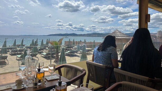 En restaurant ved havet med udendørs siddepladser med udsigt over havet, et træbord med glas og en flaske, samt udsigt til stranden og havet.