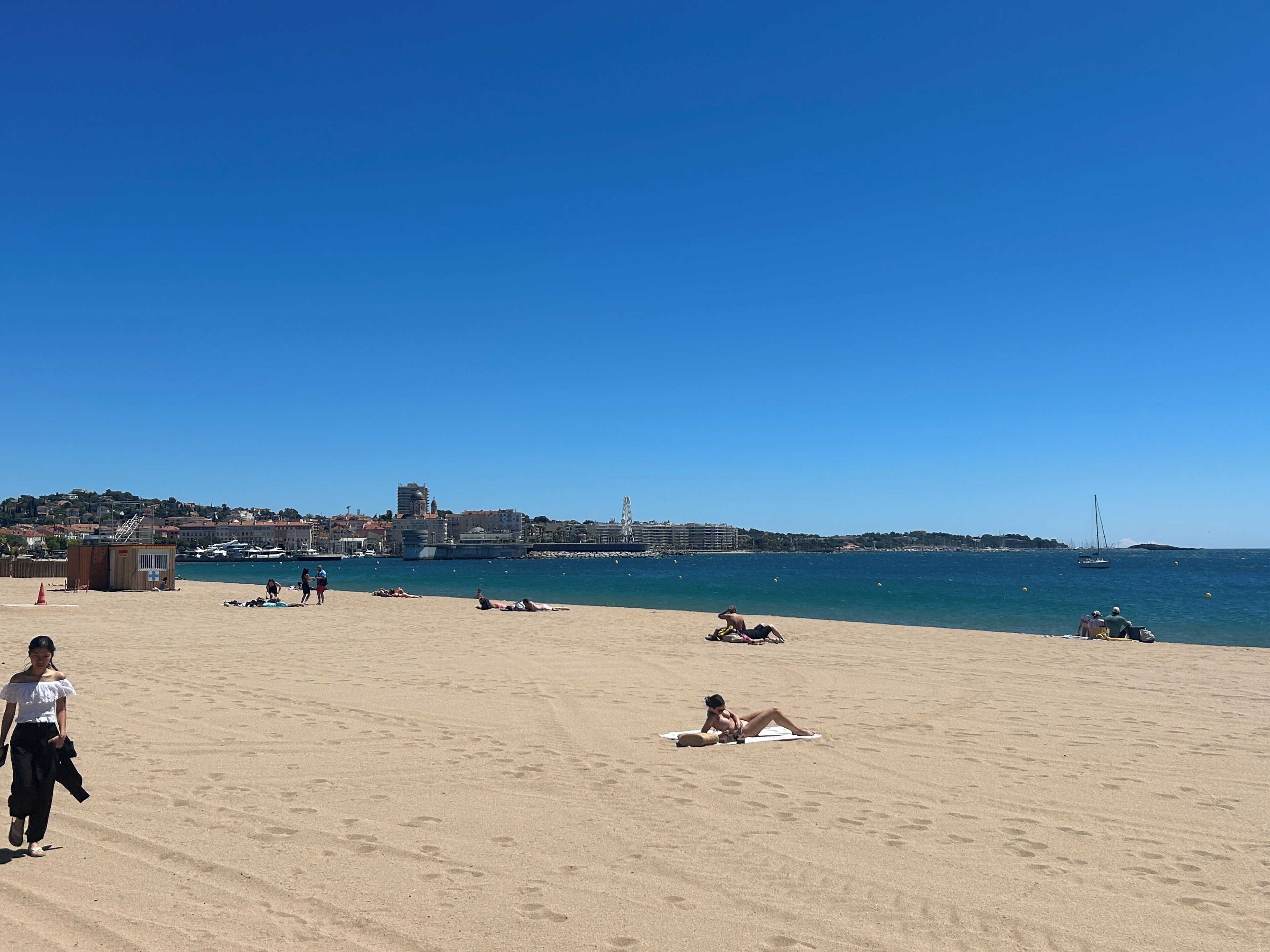A beach with people sunbathing and walking, a clear sky, and a distant cityscape.