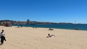 En strand med solbadere og gående mennesker, en klar himmel og en by i det fjerne.
