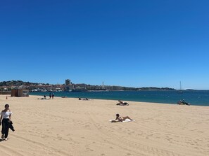 A beach with people sunbathing and walking, a clear sky, and a distant cityscape. - Bright duplex 75m2, 1 minute walk from the beach (Fréjus)