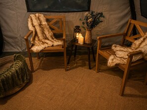A cozy interior with two wooden chairs, a woven rug, a small table with candles and a lantern, and a green pouf.