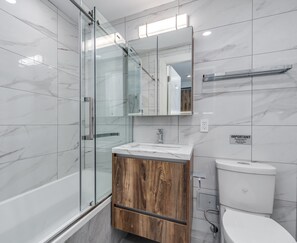 Een moderne badkamer met een douchecabine van glas, een houten wastafelmeubel, een wit toilet en een spiegel boven de wastafel.