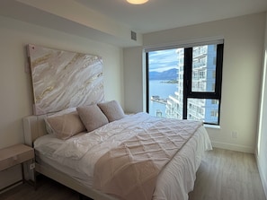 A bedroom with a bed, a nightstand, a large window with a view of a body of water and buildings, and a marble-patterned headboard. - Luxury Lakeview Condo at Aqua - Pool & Sandy Beach (Kelowna)