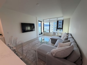 A modern living room with a flat-screen TV, a glass coffee table, a sofa, and a view of the sea through large windows. - Luxury Lakeview Condo at Aqua - Pool & Sandy Beach (Kelowna)