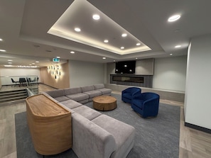 A modern lounge area with a sectional sofa, round wooden coffee table, blue armchairs, a flat-screen TV, and a fireplace. - Luxury Lakeview Condo at Aqua - Pool & Sandy Beach (Kelowna)
