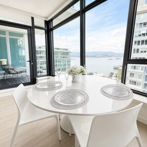 A modern dining area with a round table, white chairs, and a view of a body of water through large windows. - Luxury Lakeview Condo at Aqua - Pool & Sandy Beach (Kelowna)