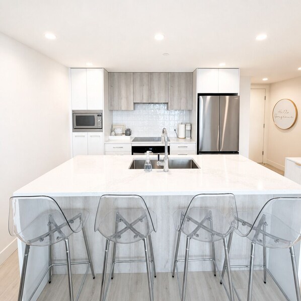 A modern kitchen with a large island, stainless steel appliances, and clear chairs. - Luxury Lakeview Condo at Aqua - Pool & Sandy Beach (Kelowna)