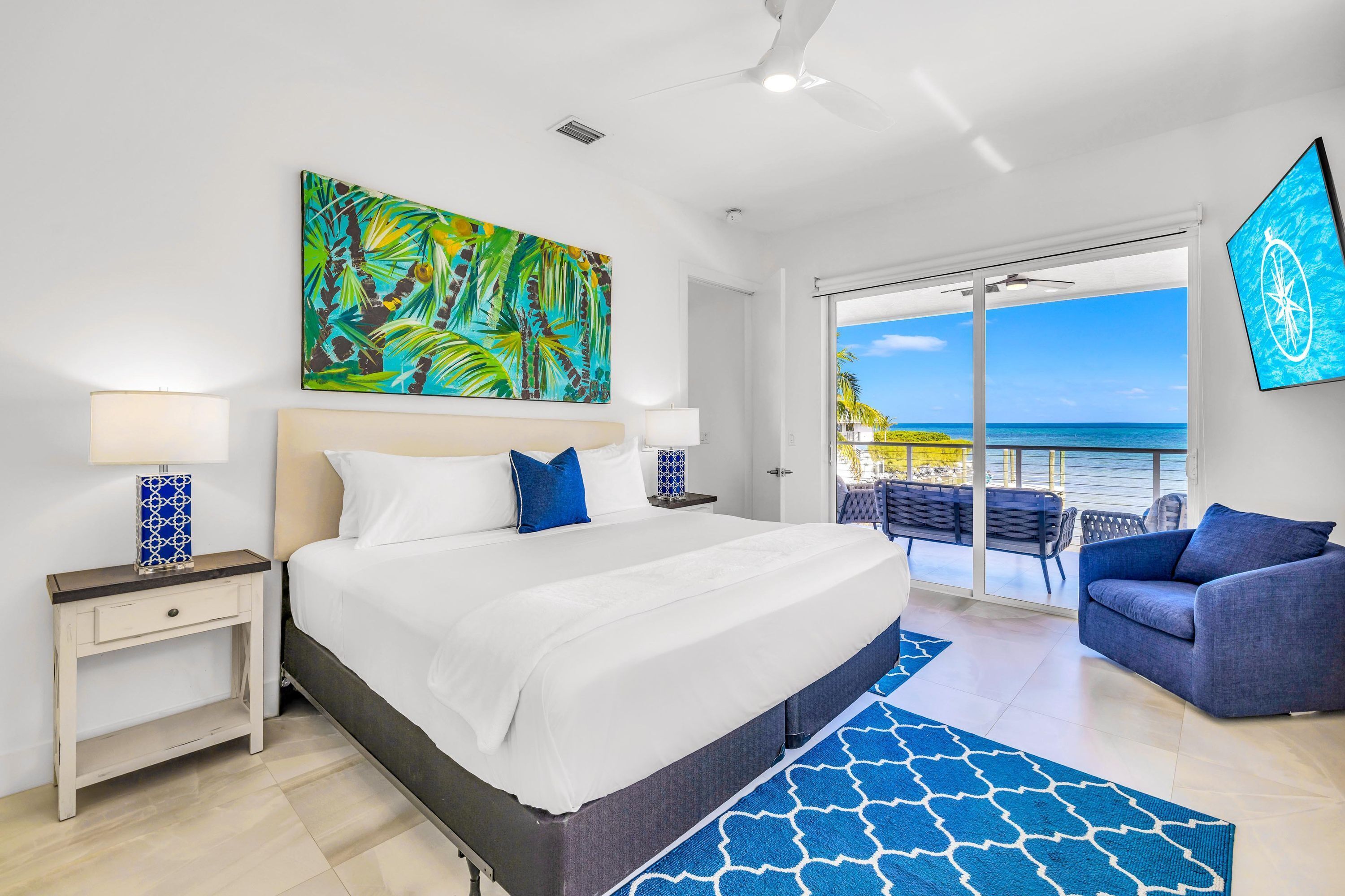 Een slaapkamer met een bed, nachtkastjes, een groot schilderij en uitzicht op zee.