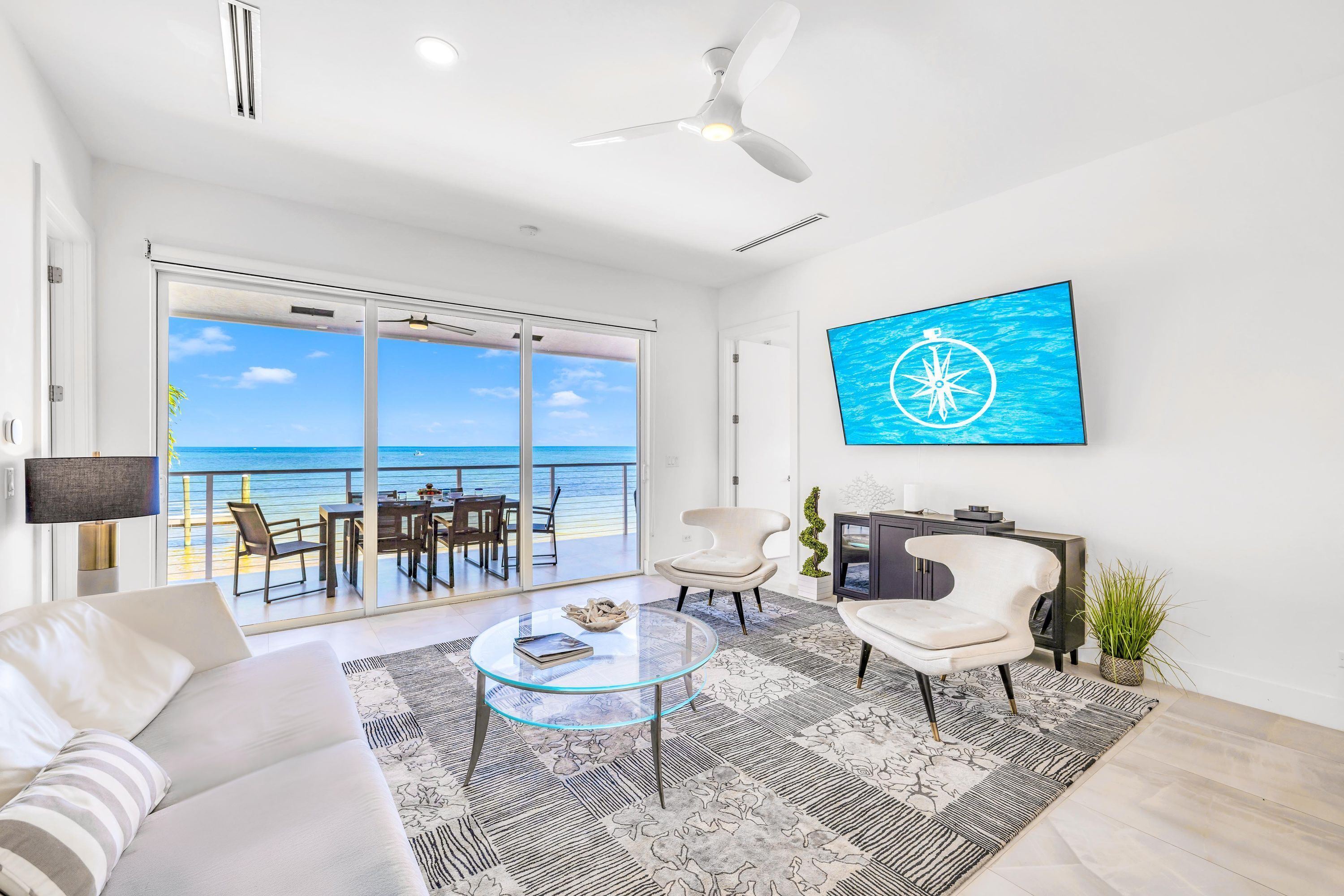 A modern living room with a glass coffee table, a sofa, a TV mounted on the wall, and a view of the ocean through large sliding doors.