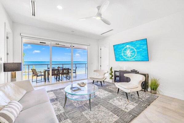 A modern living room with a glass coffee table, a sofa, a TV mounted on the wall, and a view of the ocean through large sliding doors. - Beach Residence Private Overwater Dock Heated Pool Mahi Key (Islamorada)