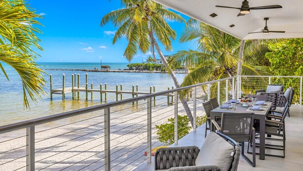 A balcony with a view of the sea, a dining area with chairs and a table, and a ceiling fan. - Beach Residence Private Overwater Dock Heated Pool Mahi Key (Islamorada)