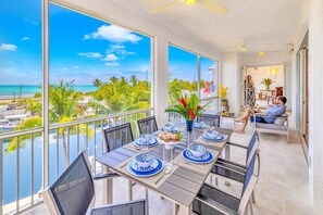 A dining area with a view of the ocean, a balcony with a railing, and a seating area with a person relaxing. - Ocean View Two Duplex Penthouses Retreat Amenities Tennis Court Heated Pool Vista Key (Islamorada)