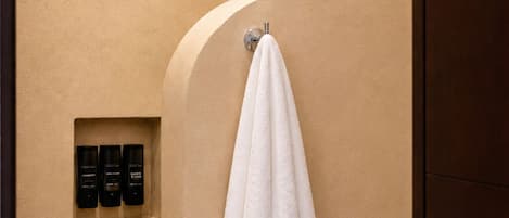 A bathroom with a shower, a towel hanging on the wall, and three bottles of toiletries in a shelf.
