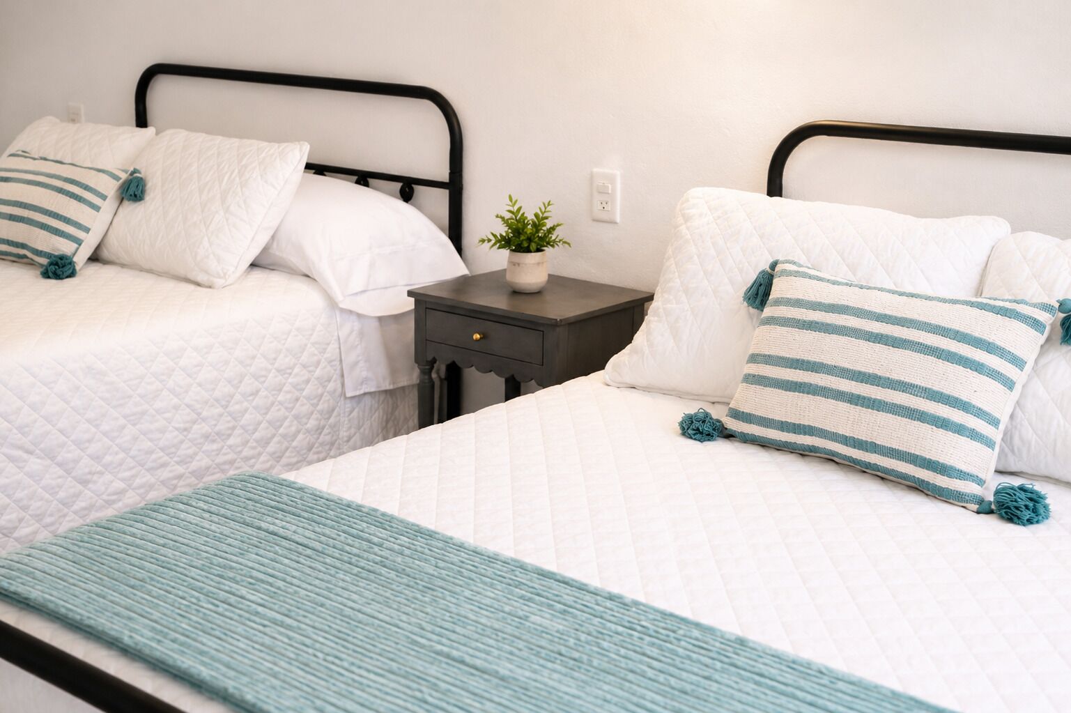 A room with two beds, each with white bedding and teal accents, a small bedside table with a plant, and a teal throw blanket.