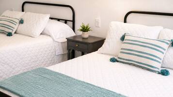 A room with two beds, each with white bedding and teal accents, a small bedside table with a plant, and a teal throw blanket.