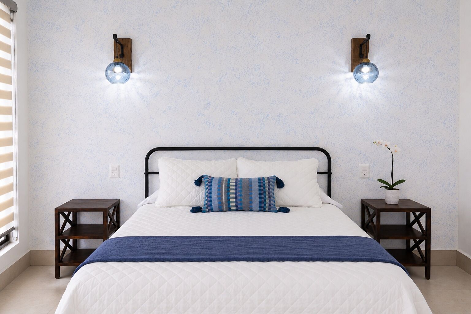 Une chambre à coucher avec un lit, deux tables de chevet, une plante en pot et des lampes fixées au mur.