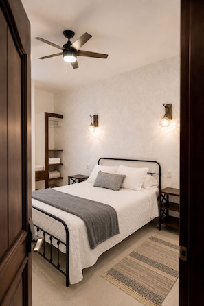 A bedroom with a bed, a ceiling fan, bedside lamps, a nightstand, a wardrobe, and a patterned rug. - Casa Feregrino Hotel Boutique (Cuatro Ciénegas de Carranza)