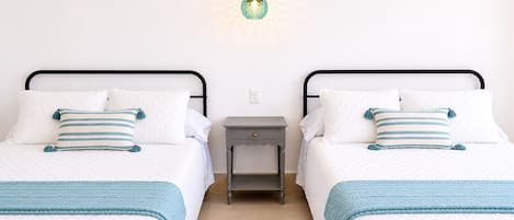 A double bed with a quilted white cover, two pillows with blue stripes, a blue throw blanket, a small bedside table, and a wall-mounted light fixture.