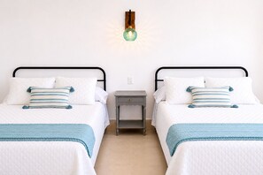 Una cama doble con cubrecolcha blanco acolchado, dos almohadas a rayas azules, una manta azul y una mesita de noche pequeña, además de una lámpara de pared.