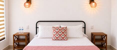 Una cama con colcha blanca y rosa, dos mesitas de noche de madera, un cabecero de madera y dos lámparas de pared.