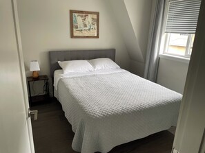 A bedroom with a bed, a bedside table with a lamp, a painting on the wall, and a window with curtains. - Cozy 2-bedroom apartment in lovely Kelowna (Kelowna)