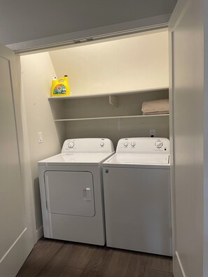 A laundry room with two white front-loading washing machines, a shelf above with a detergent bottle, and a folded towel on the top shelf. - Cozy 2-bedroom apartment in lovely Kelowna (Kelowna)