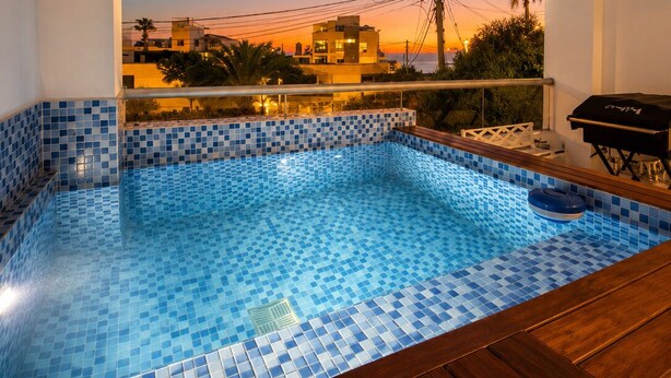 A swimming pool with blue mosaic tiles, a wooden deck, and a view of buildings at sunset. - Surf & Work Retreat - Private Pool - Fast WiFi (Punta Hermosa)