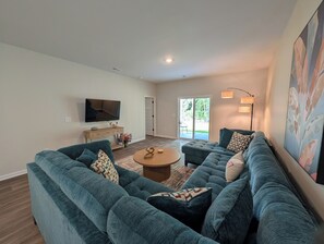 A modern living room with a sectional sofa, a coffee table, a flat-screen TV, and a sliding glass door leading to a patio. - Relax in Calabash – New 3BR/2BA Home Just Minutes to Beaches, Golf & Seafood (Calabash)