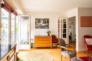 Ein Wohnzimmer mit einem Holzschrank, einem Sessel, einem roten Ledersessel, einem Gemälde an der Wand und Blick durch eine Glastür.