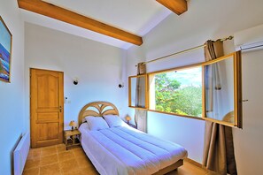 A bedroom with a bed, a wooden door, a nightstand, a lamp, a window with curtains, and a radiator. - The Terrace of the Gulf of Ajaccio - Welkeys (Pietrosella)
