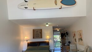 Et soveværelse med en seng, to sengeborde med lamper, en ventilator i loftet, et surfboard monteret på væggen, og udsigt udad.