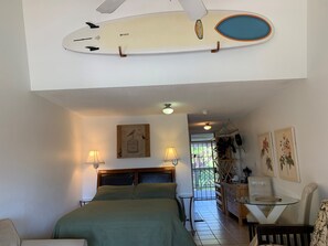 A bedroom with a bed, two bedside lamps, a ceiling fan, a wall-mounted surfboard, and a view of the outside. - Kihei Bay Surf #226 Convenient, Shared Hot Tub, Near Beach, Great Rates! (Kihei)