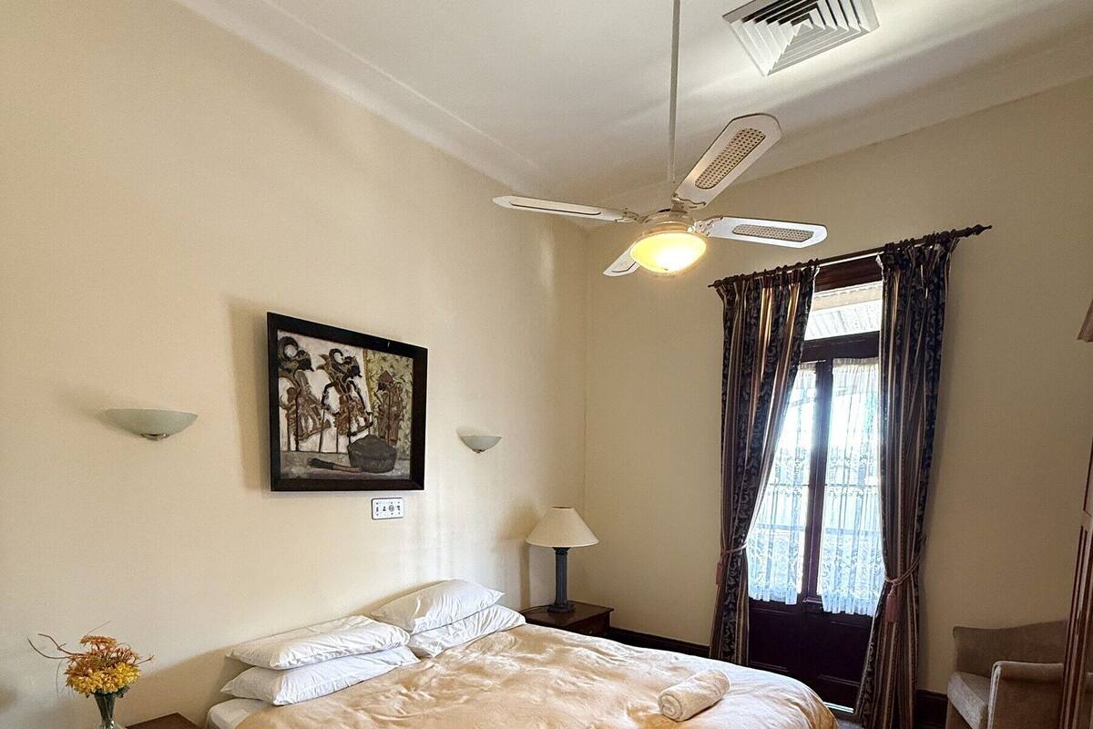 Un dormitorio con cama, ventilador de techo, ventana con cortinas, mesita de noche con un florero y un cuadro colgado en la pared.