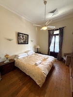 Quarto com cama, ventilador de teto, janela com cortinas, mesa de cabeceira com um vaso e um quadro pendurado na parede.
