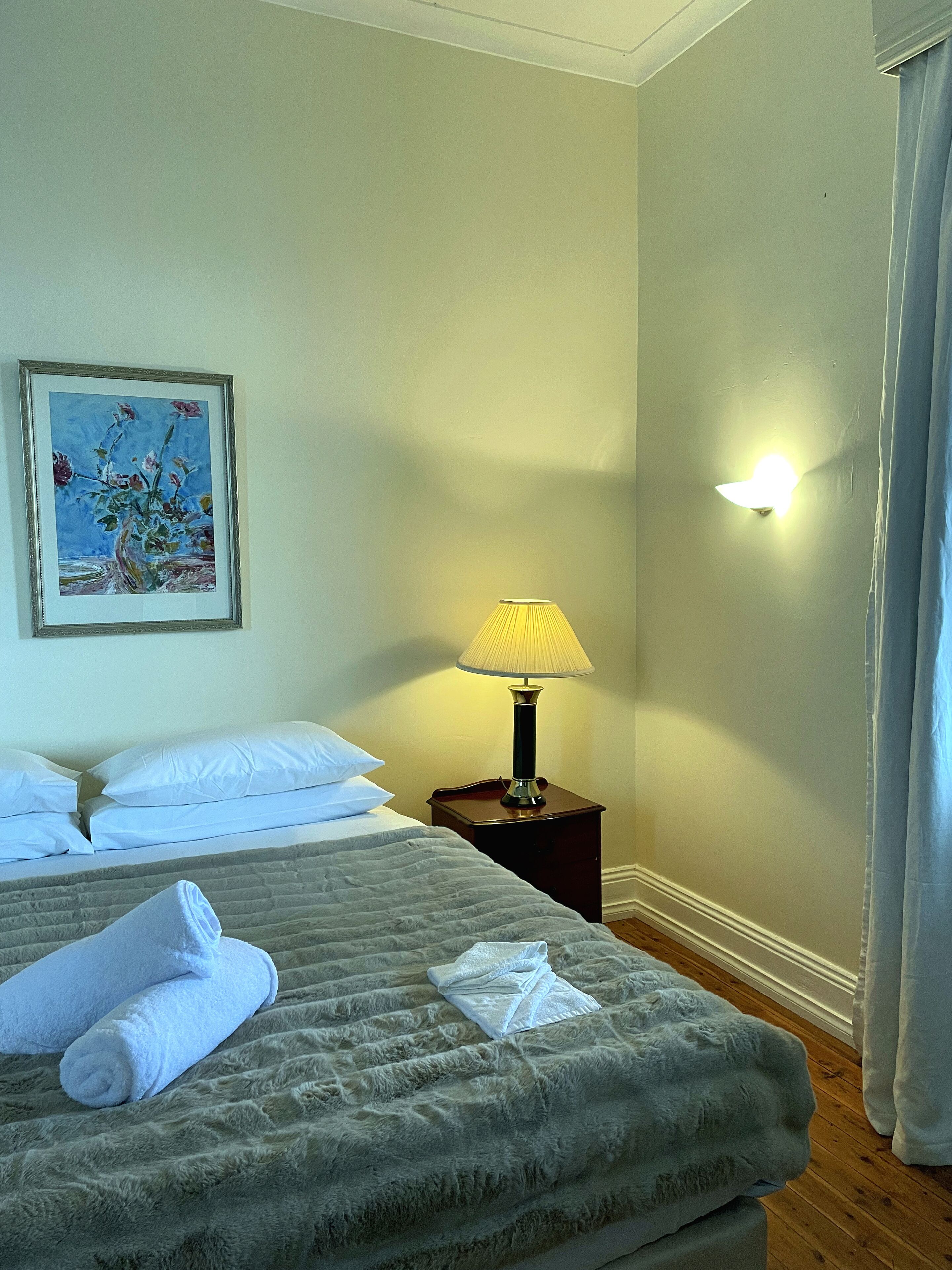 Ein Hotelzimmer mit Bett, Nachttisch mit Lampe, einem Bild an der Wand und einem Fenster mit Vorhängen.
