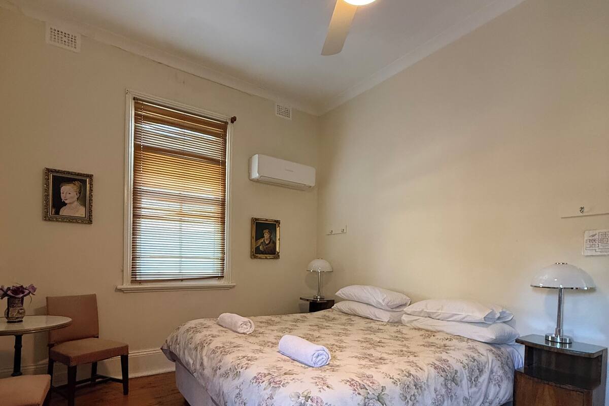 Un dormitorio con cama, ventilador de techo, ventana con persianas, mesita de noche con lámpara y una pequeña mesa con sillas.