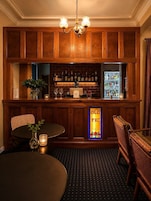 Uma área de bar com painéis de madeira, um candelabro, uma geladeira repleta de bebidas e um letreiro de vitral com a inscrição 'OLD WC'.
