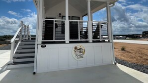 A white building with a covered porch, a staircase, and a logo on the wall. - Charming Tiny Home for five guests in Fredericksburg TX (Fredericksburg)