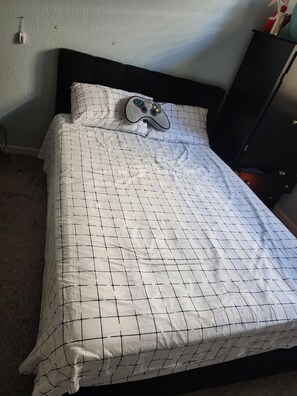 Sängyssä on valkoisella ruudukkokuvioisella päiväpeitteellä ja yhdellä tyynyllä varustettu sänky, musta pääty ja pieni yöpöytä, jonka päällä on punavalkoinen esine.
