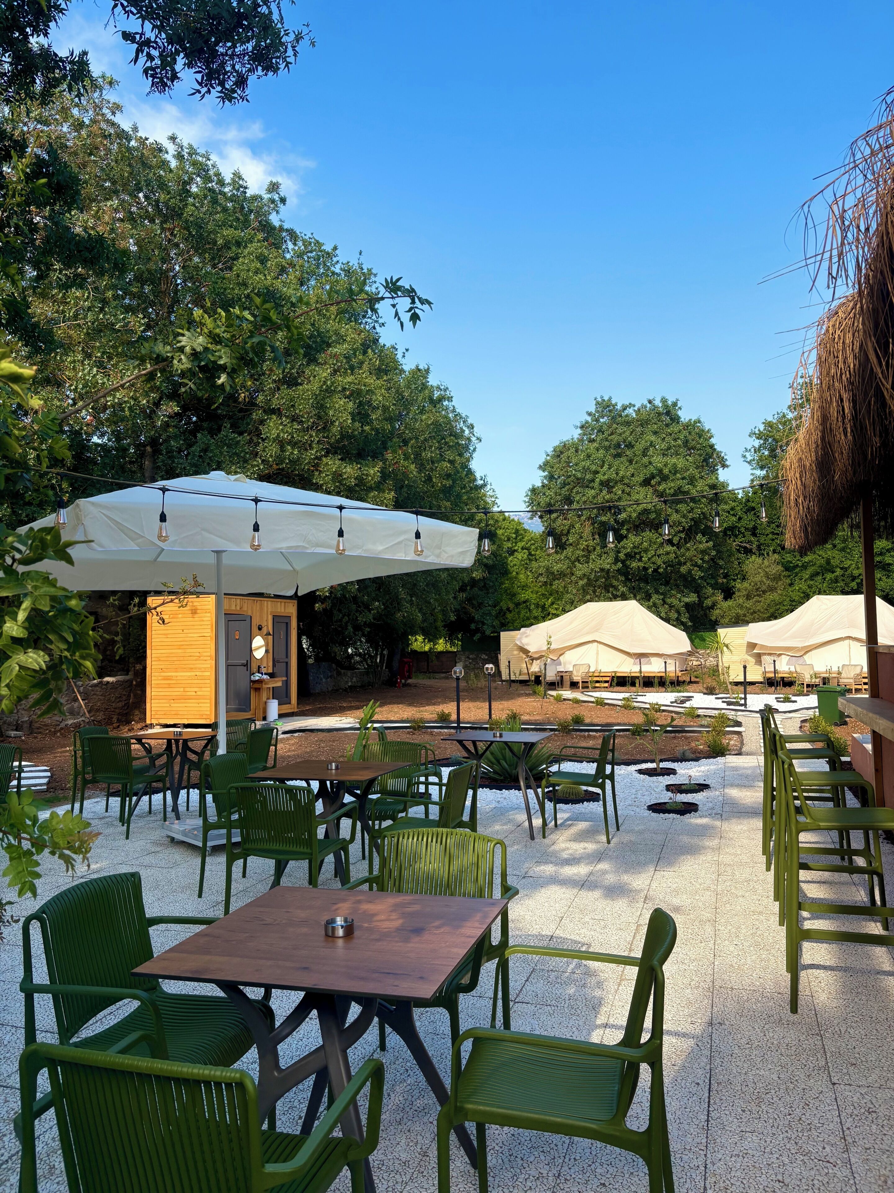 An outdoor patio with green chairs and tables, a white umbrella, and a thatched structure.