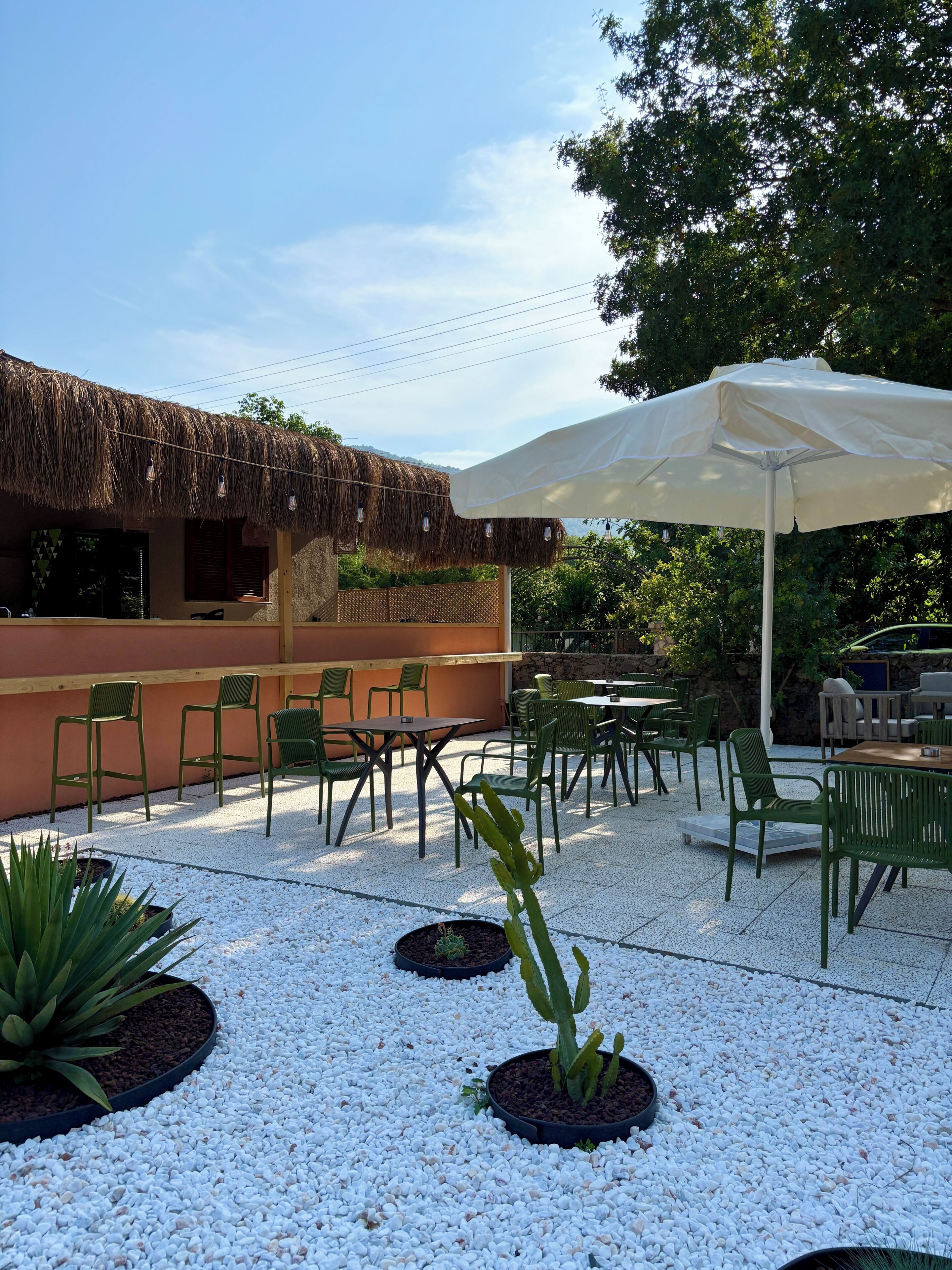 Une terrasse extérieure avec des chaises et des tables vertes, un parasol blanc et une structure à toit de chaume.