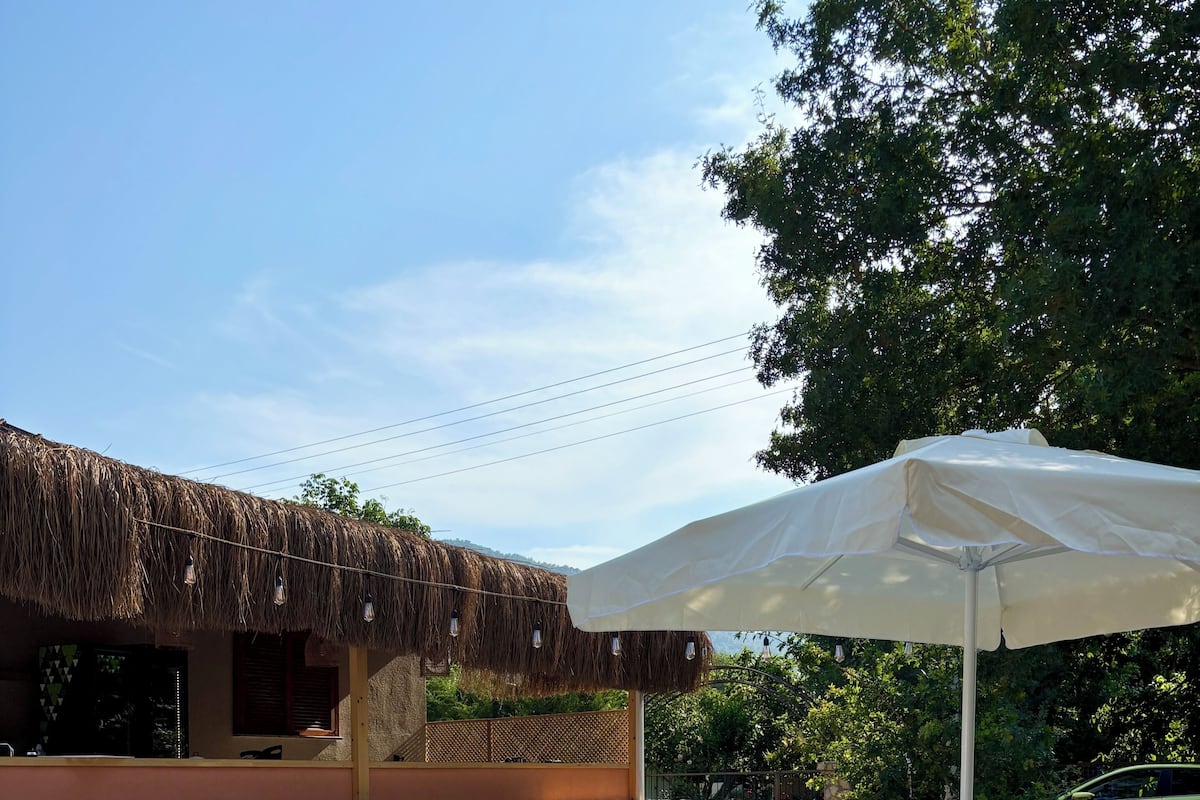 An outdoor patio with green chairs and tables, a white umbrella, and a thatched roof structure.