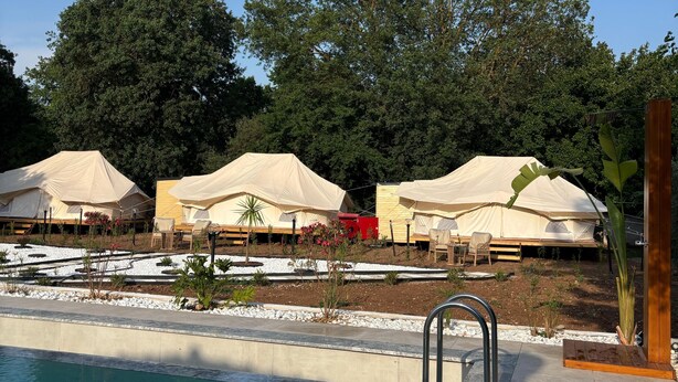 Una piscina con bordo piastrellato, circondata da una pavimentazione in cemento. Si vedono diversi tende beige con tavoli e sedie, e una struttura rossa sullo sfondo.