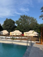 Una piscina con borde de azulejos, rodeada por un deck de hormigón. Se observan varias tiendas de campaña beige con mesas y sillas, y una estructura roja al fondo.