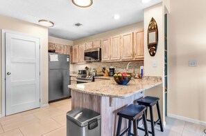 A kitchen with wooden cabinets, a granite countertop, a microwave, a stove, and a fruit bowl. - Ocean Mile K-10, Poolside views, Beach View, Beach Gear Credit with Booking (St. George Island)