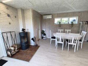 A room with a fireplace, a dining table with chairs, a window with plants, and a door. - 4 Star Holiday Home in Vaeggerlose (Vaeggerlose)
