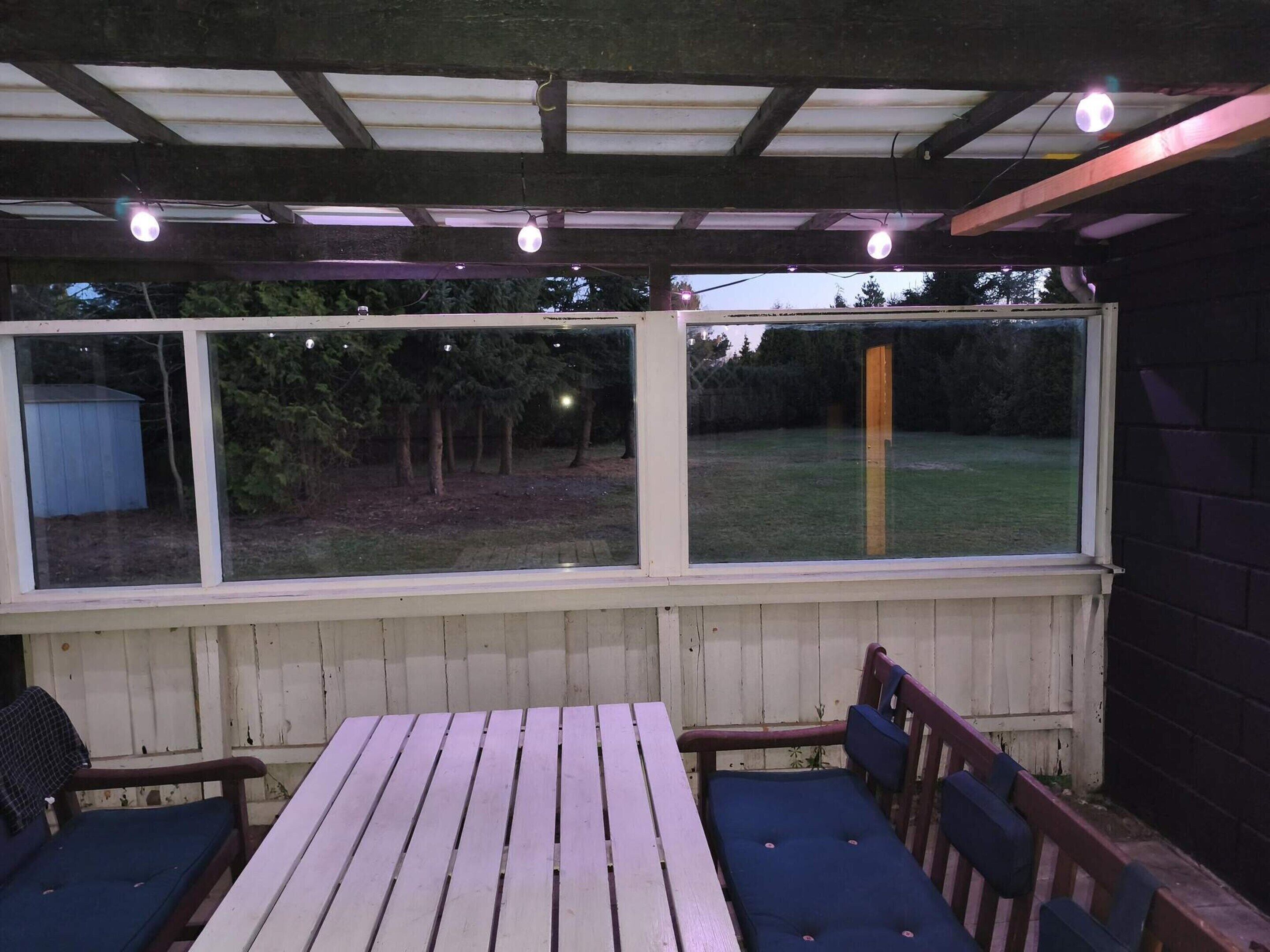Um alpendre coberto com mesa e cadeiras, janelas amplas e vista para um jardim.